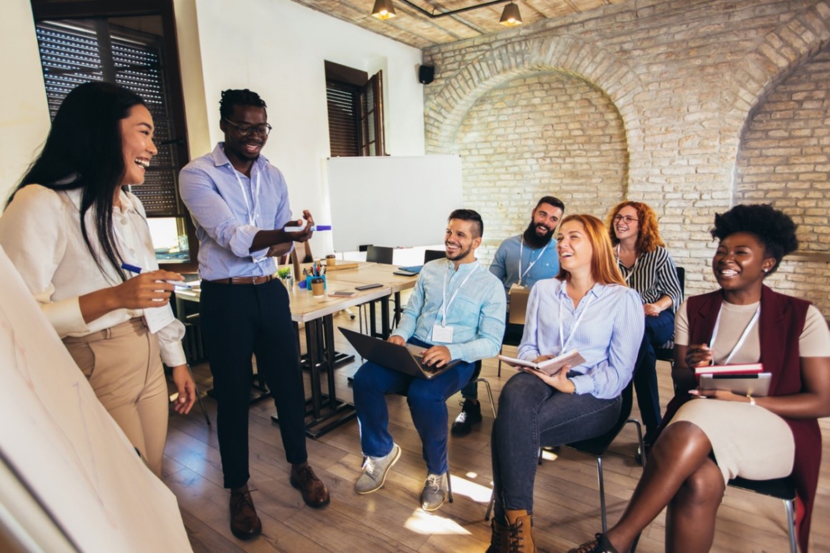 Two people in front of people in a meeting room explaining something